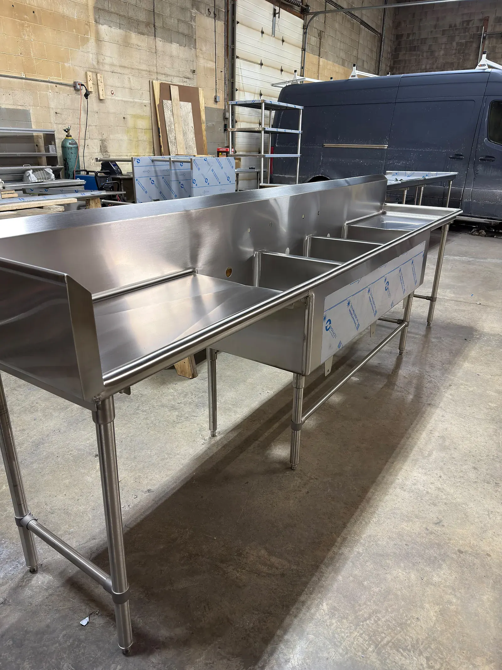 Long three compartment stainless sink with drainboards and a raised backsplash, on tubular legs in the shop