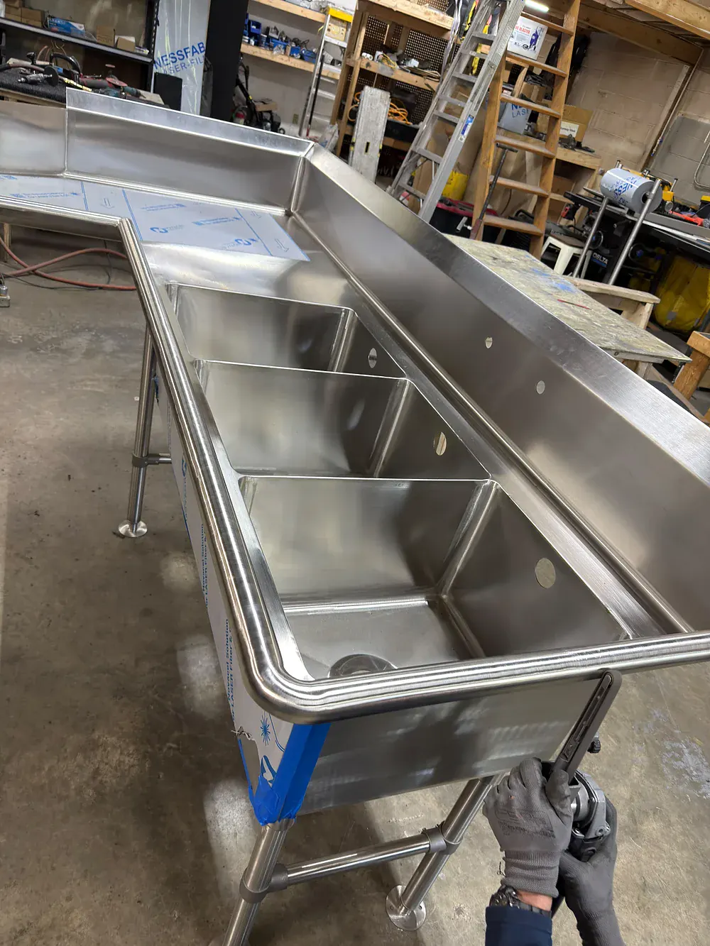 Gloved hand steadying the rim of a deep stainless sink bowl at the bench